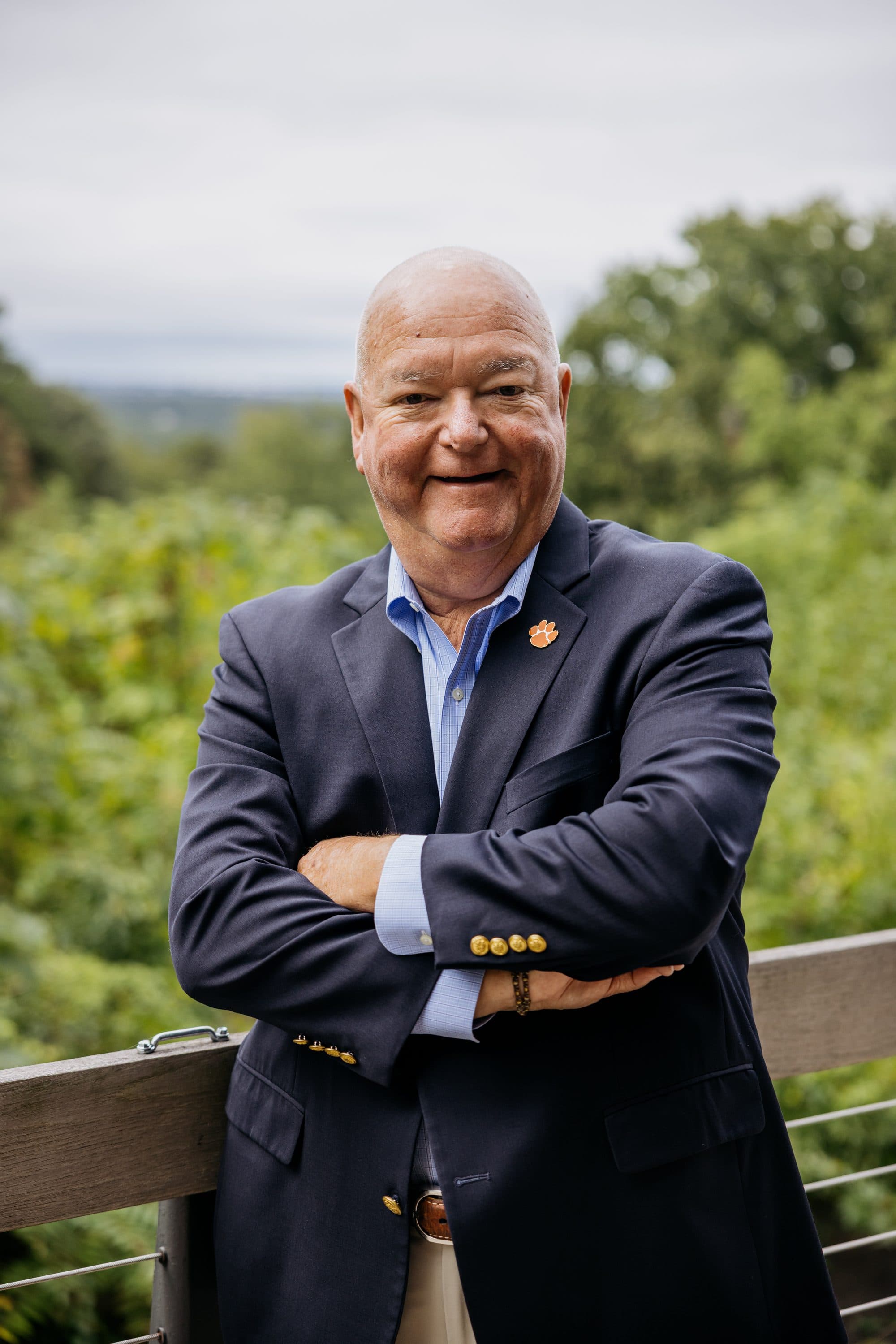 Chuck Saylors, smiling with arms crossed, leaning on an outdoor railing with trees behind him